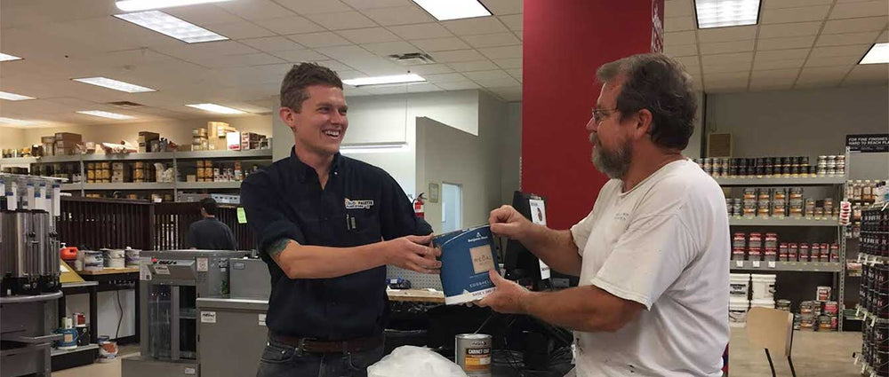 employee in black shirt handing paint can to man in white shirt inside of store