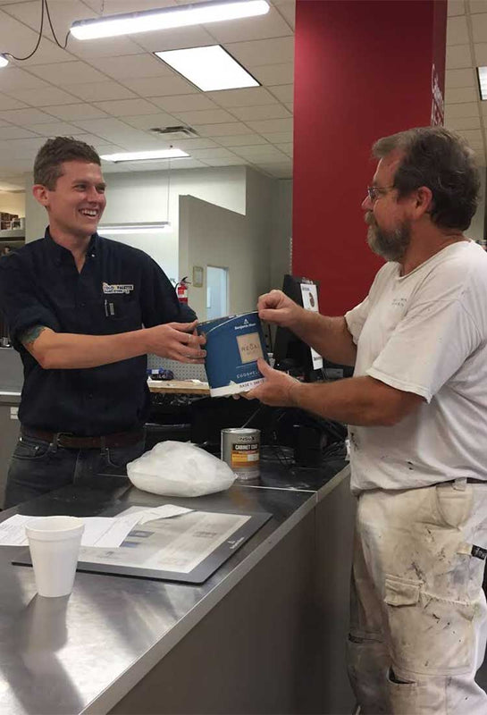 employee in black shirt handing paint can to man in white shirt inside of store